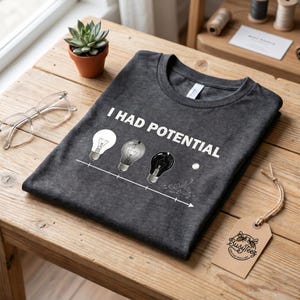 Op de afbeelding: Donkergrijs t-shirt met de tekst "I HAD POTENTIAL" en een afbeelding van drie gloeilampen, waarvan één brandt, één gedeeltelijk en één kapot. De tee is op een houten tafel gevouwen, met een kleine plant en een bril.