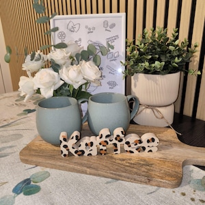 Könnte beinhalten: Zwei gesprenkelte hellblaue Keramiktassen stehen auf einem Holzbrett neben einem Holzschriftzug "Kaffee" mit Leopardenmuster. Dahinter befinden sich weiße Rosen, ein gerahmtes Nudelposter und eine kleine grüne Pflanze in einem gerippten Topf.