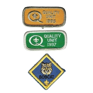May include: Three embroidered patches including a rectangular gold patch with the text QUALITY UNIT 1995, a rectangular green patch with the text QUALITY UNIT 1997, and a diamond-shaped blue patch featuring a bobcat illustration and the word BOBCAT.