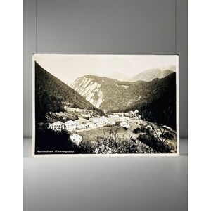 May include: A vintage black and white postcard featuring a landscape view of the village of Martinsbruck in the Unterengadin valley. The image displays houses nestled in a mountain valley with dense forests and peaks, with the text Martinsbruck (Unterengadin) printed in the bottom left corner.