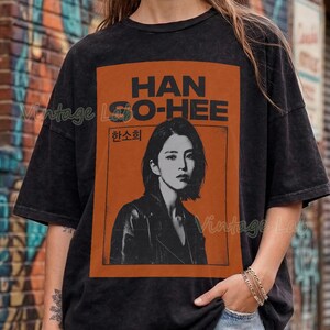Könnte beinhalten: Schwarzes T-Shirt mit einem orangefarbenen Aufdruck von Han So-hee und dem Text "HAN SO-HEE". Das Shirt hat einen Vintage-Look und wird mit Jeansshorts getragen. Der Hintergrund zeigt eine Backsteinmauer mit Graffiti.