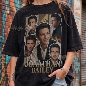 May include: Black t-shirt with a collage of Jonathan Bailey's images and the name "Jonathan Bailey" printed below. The shirt is paired with denim shorts. The background features a brick wall with graffiti art.