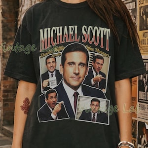 Peut inclure: T-shirt noir avec un graphisme de Michael Scott de The Office. Le design comprend le texte "MICHAEL SCOTT" et "Dunder Mifflin Office Manager" avec plusieurs images du personnage. Style vintage.
