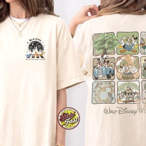 Op de afbeelding: Cr&egrave;mekleurig T-shirt met Disney-thema graphics op de voor- en achterkant. De afbeelding op de voorkant bevat personages en tekst. De achterkant toont een raster van Disney-personagesc&egrave;nes met de tekst "Walt Disney World".
