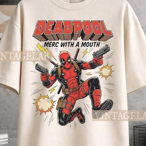Pode incluir: Uma t-shirt de cor creme com um gr&aacute;fico da personagem Deadpool num fato vermelho e preto, segurando duas pistolas. O texto "DEADPOOL" e "MERC WITH A MOUTH" est&aacute; impresso acima da personagem, acompanhado por &iacute;cones de raios amarelos.