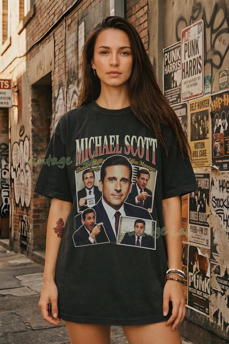 Peut inclure: T-shirt noir avec un graphisme de Michael Scott de The Office. Le design comprend le texte "MICHAEL SCOTT" et "Dunder Mifflin Office Manager" avec plusieurs images du personnage. Style vintage.