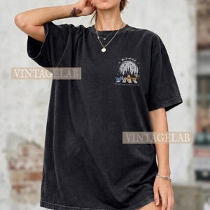 Op de afbeelding: Zwart T-shirt met een grafische print van een nachtelijke bossc&egrave;ne met cartoonfiguren en de tekst "The most magical place on earth". Het shirt heeft korte mouwen en een relaxte pasvorm.