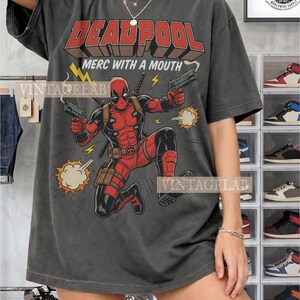 Pode incluir: Uma t-shirt cinza-carv&atilde;o com uma estampa gr&aacute;fica da personagem Deadpool em um traje vermelho e preto, segurando duas pistolas. O texto diz DEADPOOL e MERC WITH A MOUTH, acompanhado por raios amarelos e ilustra&ccedil;&otilde;es de nuvens de fuma&ccedil;a.