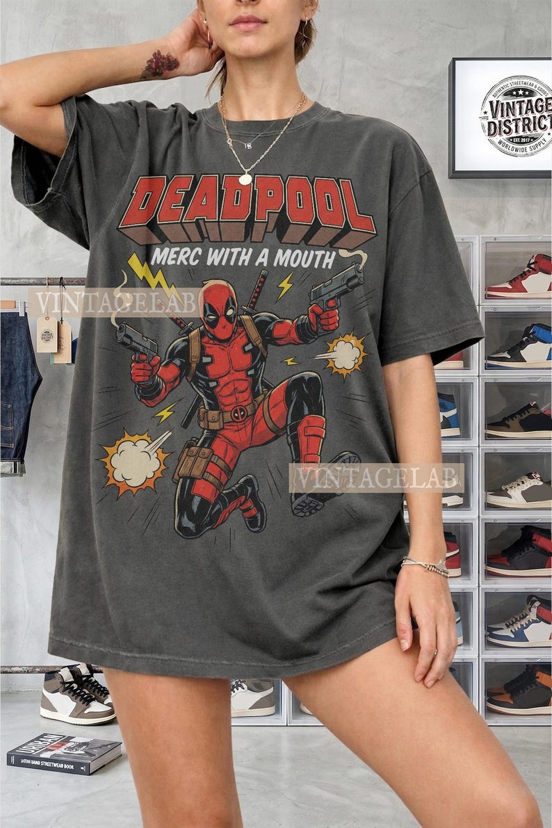 Pode incluir: Uma t-shirt cinza-carv&atilde;o com uma estampa gr&aacute;fica da personagem Deadpool em um traje vermelho e preto, segurando duas pistolas. O texto diz DEADPOOL e MERC WITH A MOUTH, acompanhado por raios amarelos e ilustra&ccedil;&otilde;es de nuvens de fuma&ccedil;a.