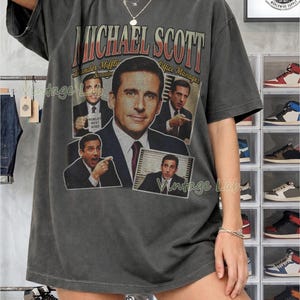 Peut inclure: T-shirt gris fonc&eacute; avec un graphisme de Michael Scott de The Office. Le design comprend plusieurs images de Scott, le texte "MICHAEL SCOTT" et "Dunder Mifflin Scranton Office Manager". Le t-shirt a un aspect vintage.