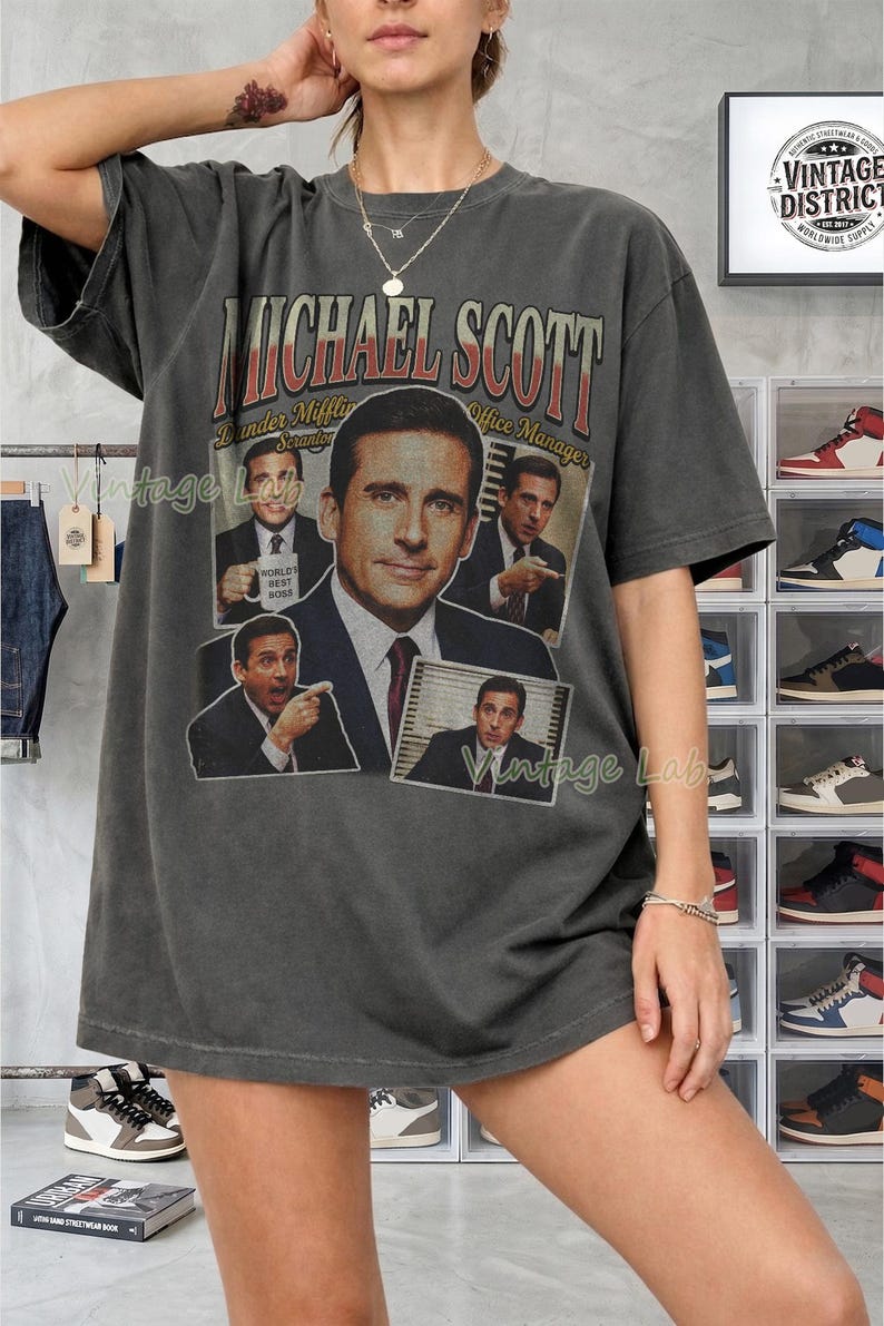 Peut inclure: T-shirt gris fonc&eacute; avec un graphisme de Michael Scott de The Office. Le design comprend plusieurs images de Scott, le texte "MICHAEL SCOTT" et "Dunder Mifflin Scranton Office Manager". Le t-shirt a un aspect vintage.