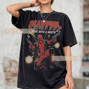 Pode incluir: Uma t-shirt preta de manga curta com um gr&aacute;fico da personagem Deadpool em fato vermelho e preto a segurar duas pistolas. O texto DEADPOOL e MERC WITH A MOUTH est&aacute; impresso acima da personagem com rel&acirc;mpagos amarelos e nuvens de fumo.