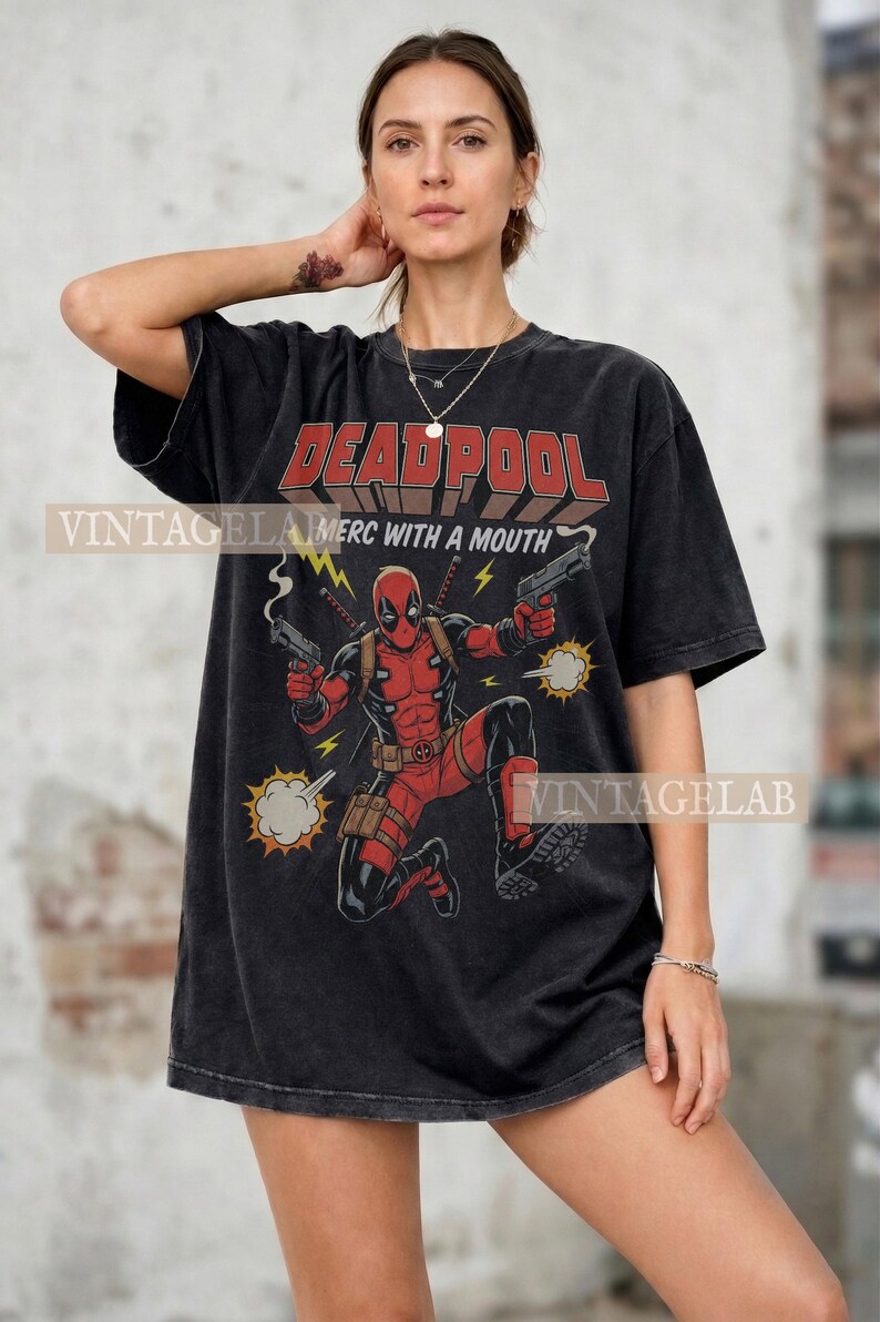 Pode incluir: Uma t-shirt preta de manga curta com um gr&aacute;fico da personagem Deadpool em fato vermelho e preto a segurar duas pistolas. O texto DEADPOOL e MERC WITH A MOUTH est&aacute; impresso acima da personagem com rel&acirc;mpagos amarelos e nuvens de fumo.
