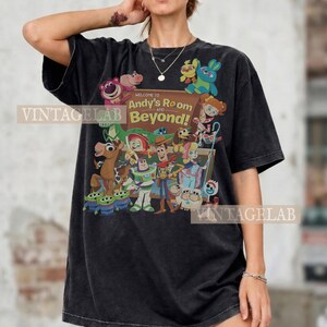 K&ouml;nnte beinhalten: Schwarzes T-Shirt mit einer farbenfrohen Grafik von Charakteren aus dem Film "Toy Story". Das Design enth&auml;lt den Text "Welcome to Andy's Room and Beyond!". Das Shirt hat eine lockere Passform und kurze &Auml;rmel.