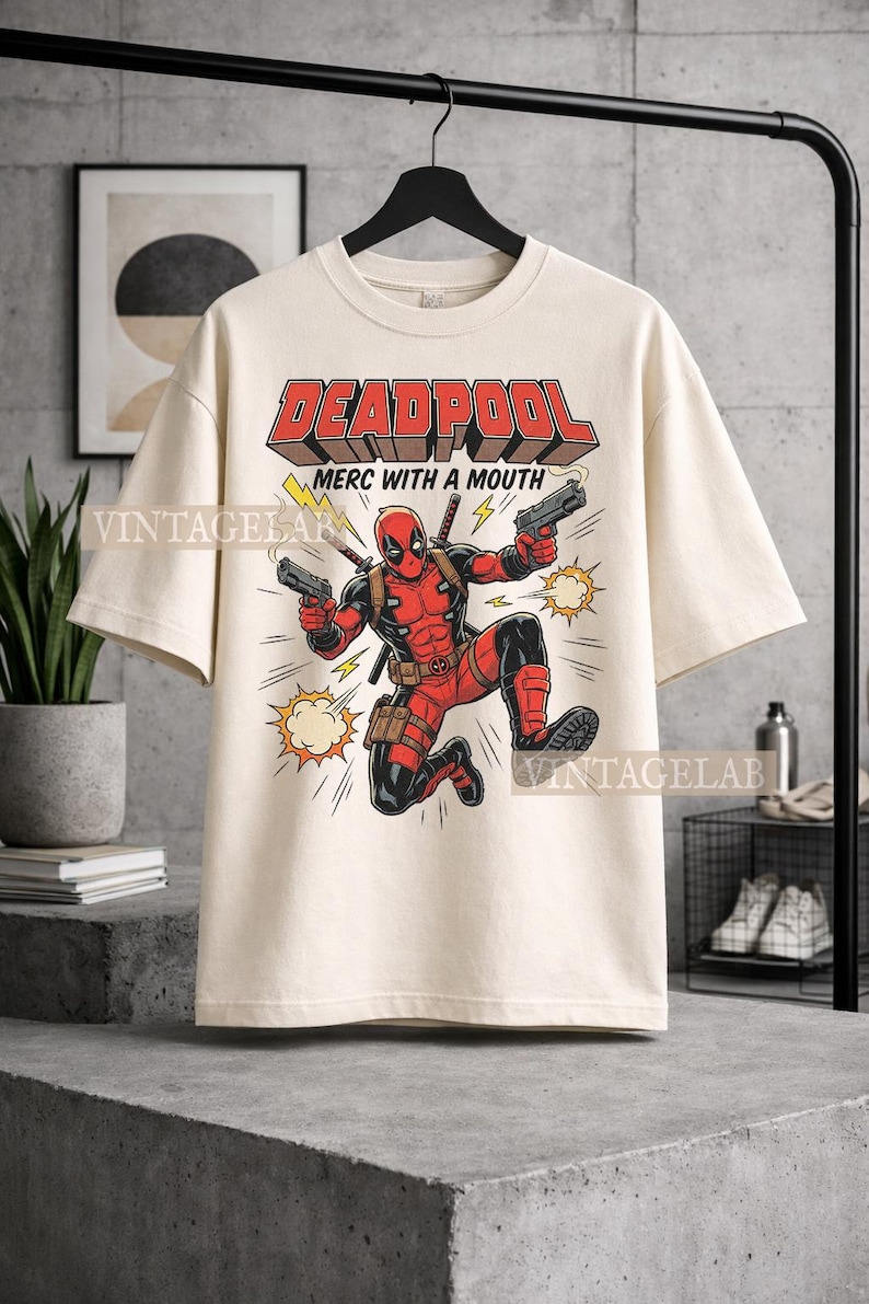 Pode incluir: Uma t-shirt de cor creme com um gr&aacute;fico da personagem Deadpool num fato vermelho e preto, segurando duas pistolas. O texto "DEADPOOL" e "MERC WITH A MOUTH" est&aacute; impresso acima da personagem, acompanhado por &iacute;cones de raios amarelos.