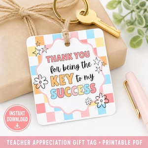 May include: A white gift tag with the text "THANK YOU for being the KEY to my SUCCESS" in colorful letters. The tag has a floral and star design, and is attached to a gold key and keychain. The tag is a teacher appreciation gift.