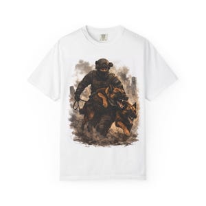 Può includere: T-shirt bianca con un'illustrazione dettagliata di una persona in equipaggiamento tattico con due pastori tedeschi. L'opera raffigura un ambiente urbano fumoso. La maglietta ha un girocollo e maniche corte.