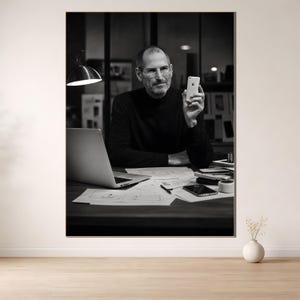 Peut inclure: Photographie en noir et blanc d'un homme tenant un smartphone, assis à un bureau avec un ordinateur portable et des croquis de conception. L'homme porte un col roulé noir. Une lampe de bureau éclaire l'espace de travail.