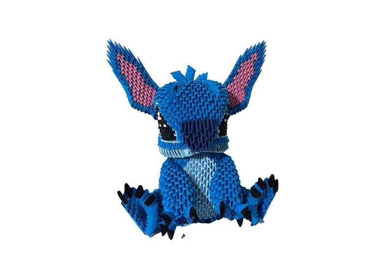May include: A blue origami figure of a cartoon character with large ears and black eyes. The ears have pink interiors. The figure is sitting with its arms and legs outstretched. The figure is made of many small, folded pieces of paper.