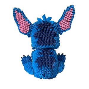 May include: A blue origami figure of the character Stitch from Lilo & Stitch, with pink inner ears and black claws. The figure is made of folded paper and sits on a white background.