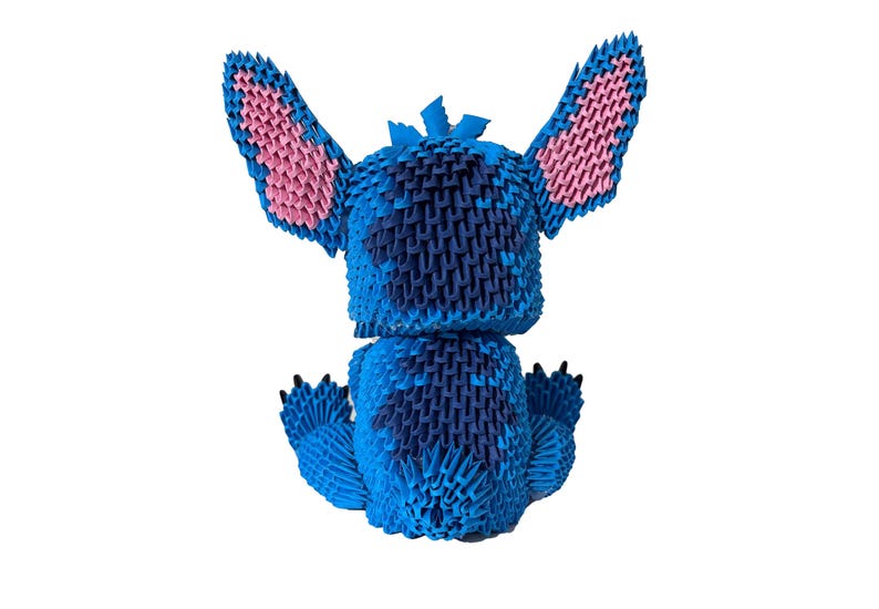 May include: A blue origami figure of the character Stitch from Lilo & Stitch, with pink inner ears and black claws. The figure is made of folded paper and sits on a white background.