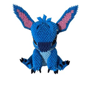 May include: A blue origami figure of a cartoon character with large ears and black eyes. The ears have pink inner details. The figure is sitting with its arms and legs spread out. The origami is made of many small, folded paper pieces.