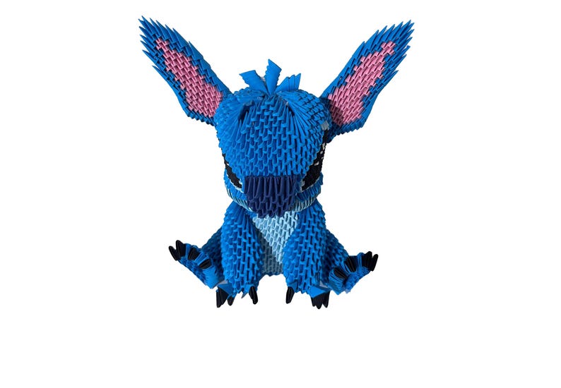 May include: A blue origami figure of a cartoon character with large ears and black eyes. The ears have pink inner details. The figure is sitting with its arms and legs spread out. The origami is made of many small, folded paper pieces.