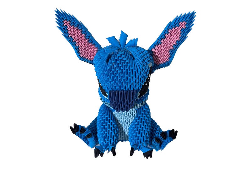 May include: A blue origami figure of a cartoon character with large ears and black eyes. The ears have pink inner details. The figure is sitting with its arms outstretched, and its feet are black.