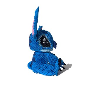 May include: A blue origami figure of a cartoon character with large black eyes, a wide mouth, and large ears. The figure is sitting down, with its arms and legs folded. The figure is made of many small, folded pieces of paper.