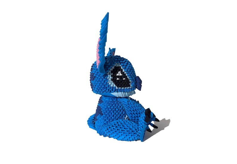 May include: A blue origami figure of a cartoon character with large black eyes, a wide mouth, and large ears. The figure is sitting down, with its arms and legs folded. The figure is made of many small, folded pieces of paper.