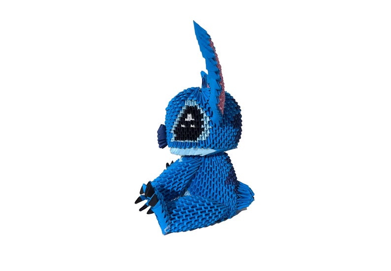 May include: A blue origami figure of a cartoon character with large ears and black eyes. The figure is sitting down with its arms and legs in front. The origami is made of many small, folded pieces of paper.