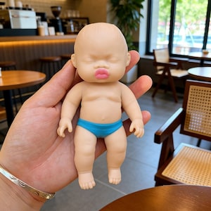 May include: A small baby doll with a realistic appearance, featuring closed eyes and a pout. The doll has a light skin tone, pink lips, and is wearing blue briefs. It's a miniature representation of a baby.