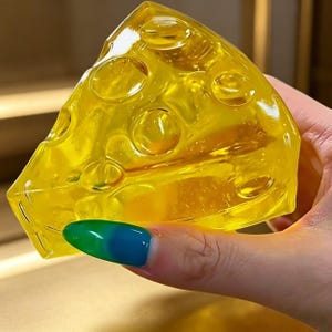 May include: A wedge of yellow, translucent cheese with several round holes. The cheese is held in a hand with a painted nail. The cheese appears to be made of a solid material, possibly resin or plastic.