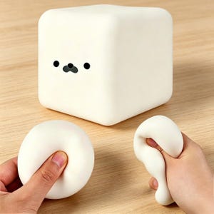 Puede incluir: Una luz nocturna blanca en forma de cubo con una cara de foca de dibujos animados. También hay dos bolas antiestrés blancas y blandas en la imagen. Los objetos están sobre una superficie de madera de color marrón claro.