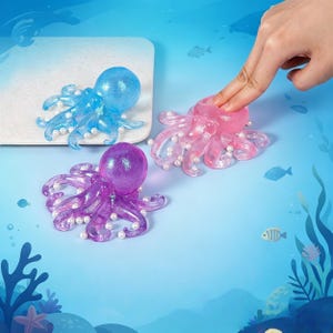 以下が含まれることがあります： 青、ピンク、紫の3色のキラキラ光るタコの置物。それぞれ小さな白いビーズで飾られています。タコの置物は、水中をテーマにした青色の背景に配置されています。