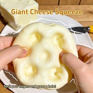 Puede incluir: Un juguete blando en forma de queso amarillo claro que se presiona con los dedos, creando agujeros. El texto "Giant Cheese Squeeze" y "Suck your fingers and press a hole!" son visibles. Un bloque de queso y un cuchillo están en el fondo.