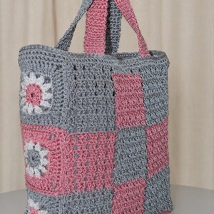 Peut inclure: Un sac fourre-tout en crochet fait main avec un motif en damier alternant des carrés gris et roses. Le panneau latéral présente trois motifs floraux avec des pétales blancs et des centres roses. Le sac comprend deux anses en crochet assorties.