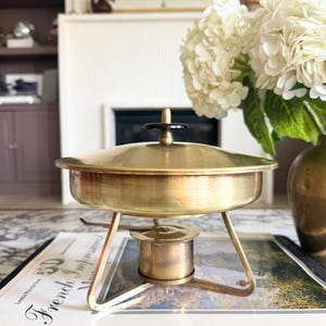 May include: A vintage brass chafing dish with a lid and a small burner. The dish has a round base supported by three legs. The lid has a black handle. The chafing dish is sitting on a book with the words "French Impressions".