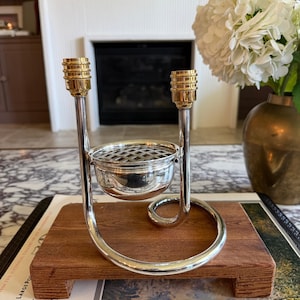 May include: A silver-toned metal flower frog stand featuring a central bowl with a grid top, flanked by two vertical rods topped with gold-toned ribbed candle holders. The structure is supported by a curved base resting on a rectangular wooden block.