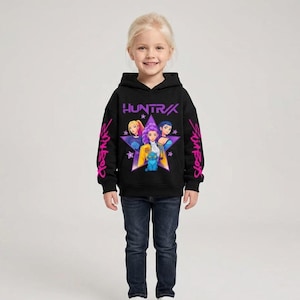 May include: Black hooded sweatshirt featuring a graphic of three characters inside a purple star with the word "HUNTRAX" above them. The sleeves have pink text. The sweatshirt is worn with blue jeans.