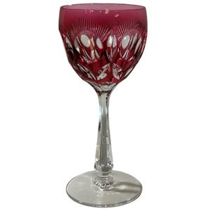 May include: A ruby red crystal wine glass with intricate cut patterns. The glass features a long, clear stem with a decorative base and a wide, bowl-shaped top. The cut design creates a starburst effect.
