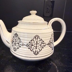 Peut inclure: Une théière en céramique de couleur crème avec une texture côtelée horizontale et un motif damassé brun foncé répété autour du centre. La théière comprend un couvercle assorti avec un petit bouton, un bec verseur courbé et une anse dorée.