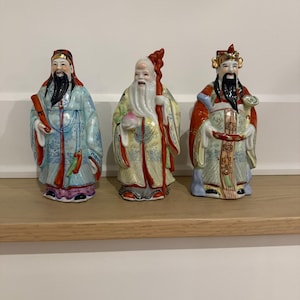 Peut inclure: Trois figurines en porcelaine représentant des personnages en robes traditionnelles. La figurine de gauche porte du bleu clair, celle du centre en jaune tient une pêche et un bâton, et celle de droite porte du rouge et de l'or. Détails peints.