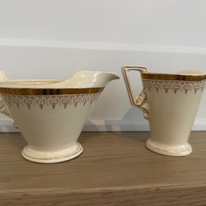 May include: Cream-colored vintage creamer and sugar bowl set. Both pieces feature gold trim with a decorative pattern. The creamer has a spout and handle, while the sugar bowl has a handle. The set is displayed on a wooden surface.