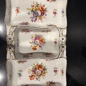 Könnte beinhalten: Eine weiße Keramik-Servierschale mit drei Fächern, die in der Mitte jedes Abschnitts mit bunten Blumensträußen verziert ist. Die Ränder sind mit Goldfarbe abgesetzt und die Seiten zeigen goldfarbene, geformte Schleifendetails.