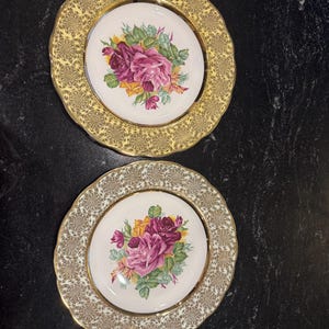 Peut inclure: Deux assiettes rondes en céramique à bords festonnés. L'assiette supérieure présente une bordure jaune avec un filigrane doré et un bouquet floral central de roses roses et jaunes. L'assiette inférieure a une bordure argentée avec un filigrane doré.