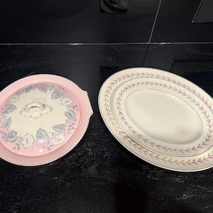 May include: A round, pink-rimmed covered dish with a floral pattern on the lid and a matching oval serving platter featuring a delicate pink and grey leaf border design. Both pieces are ceramic with cream-coloured centres and gold-toned trim.