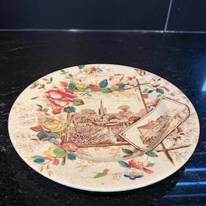 Può includere: Un piatto rotondo in ceramica con base color crema, caratterizzato da un'illustrazione centrale color seppia marrone raffigurante una cattedrale e un paesaggio. Il bordo è decorato con motivi floreali colorati in rosa, giallo e verde, incluso un piccolo riquadro rettangolare che mostra un edificio.