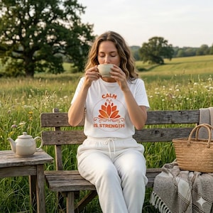Pode incluir: Uma camiseta branca com o texto "CALM IS STRENGTH" em laranja, com um desenho de flor de lótus. A pessoa está sentada em um banco de madeira, segurando uma xícara de chá. Uma chaleira e uma cesta de vime também são visíveis.