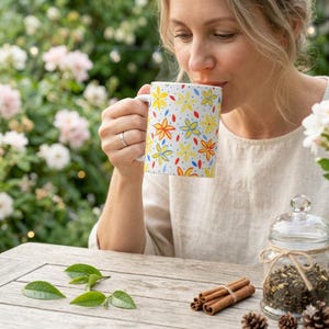 Pode incluir: Uma caneca de cerâmica branca com um padrão floral em amarelo, vermelho e azul é segurada por uma pessoa. A caneca contém uma bebida quente. A cena é ao ar livre com uma mesa de madeira, paus de canela e um frasco de vidro.